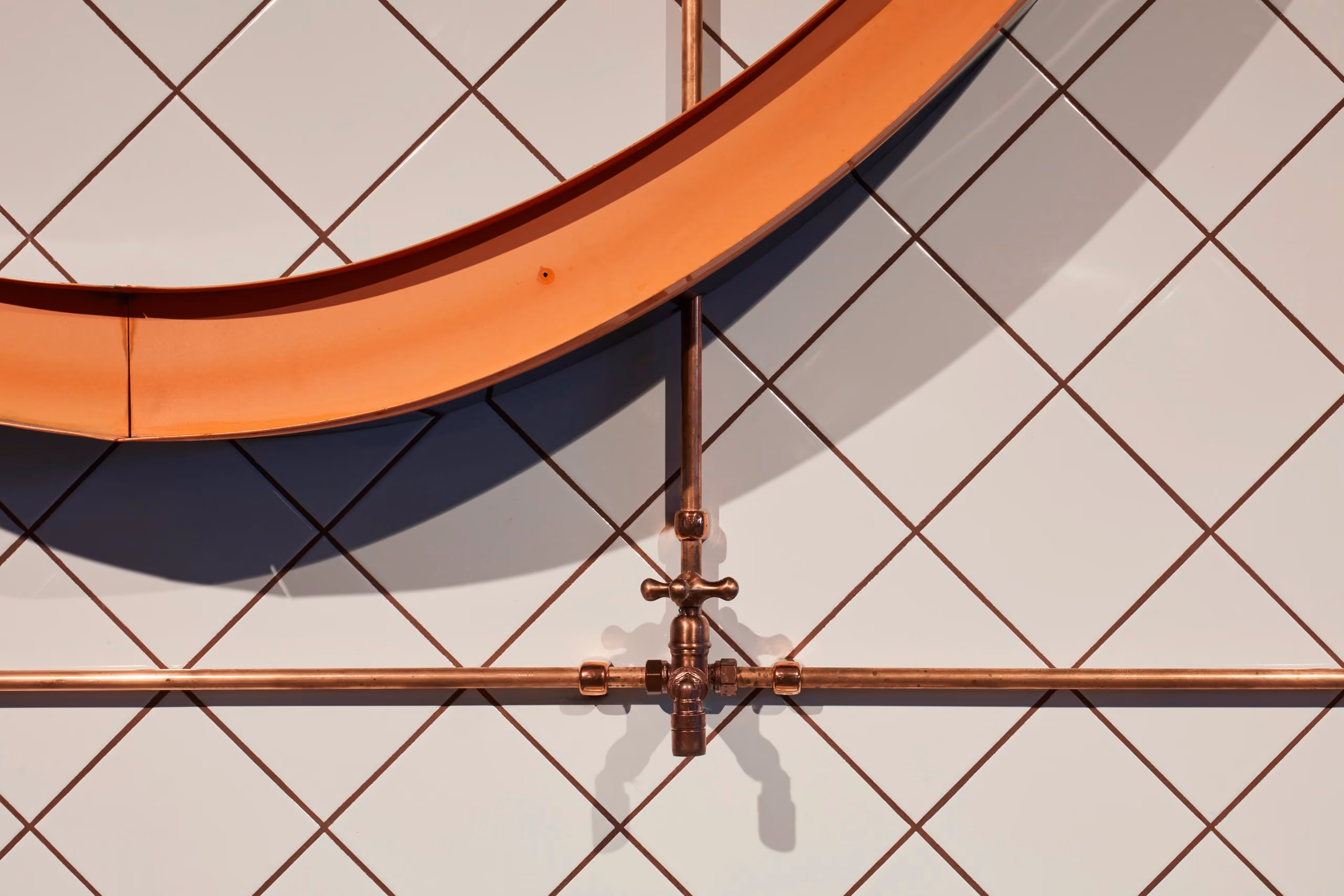 Close-up of copper pipework, warm-lit lettering and diagonal joints in the tiled drinking-water station.
