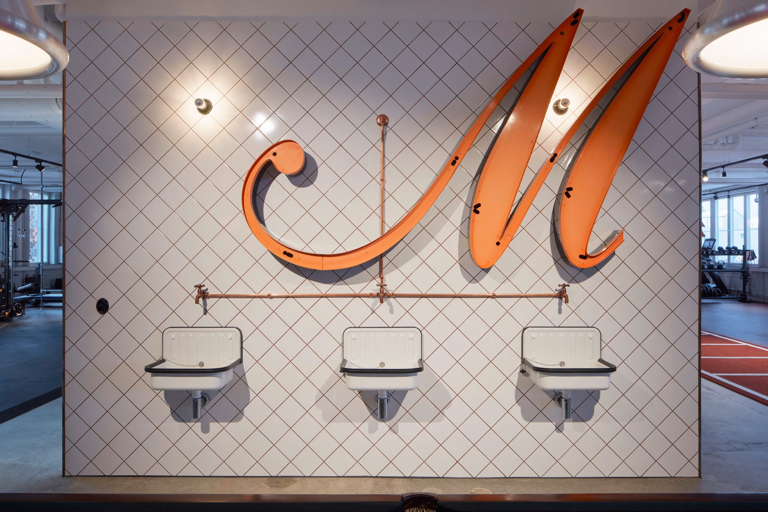 A large illuminated Mornington M above three drinking fountains on a white tiled wall.