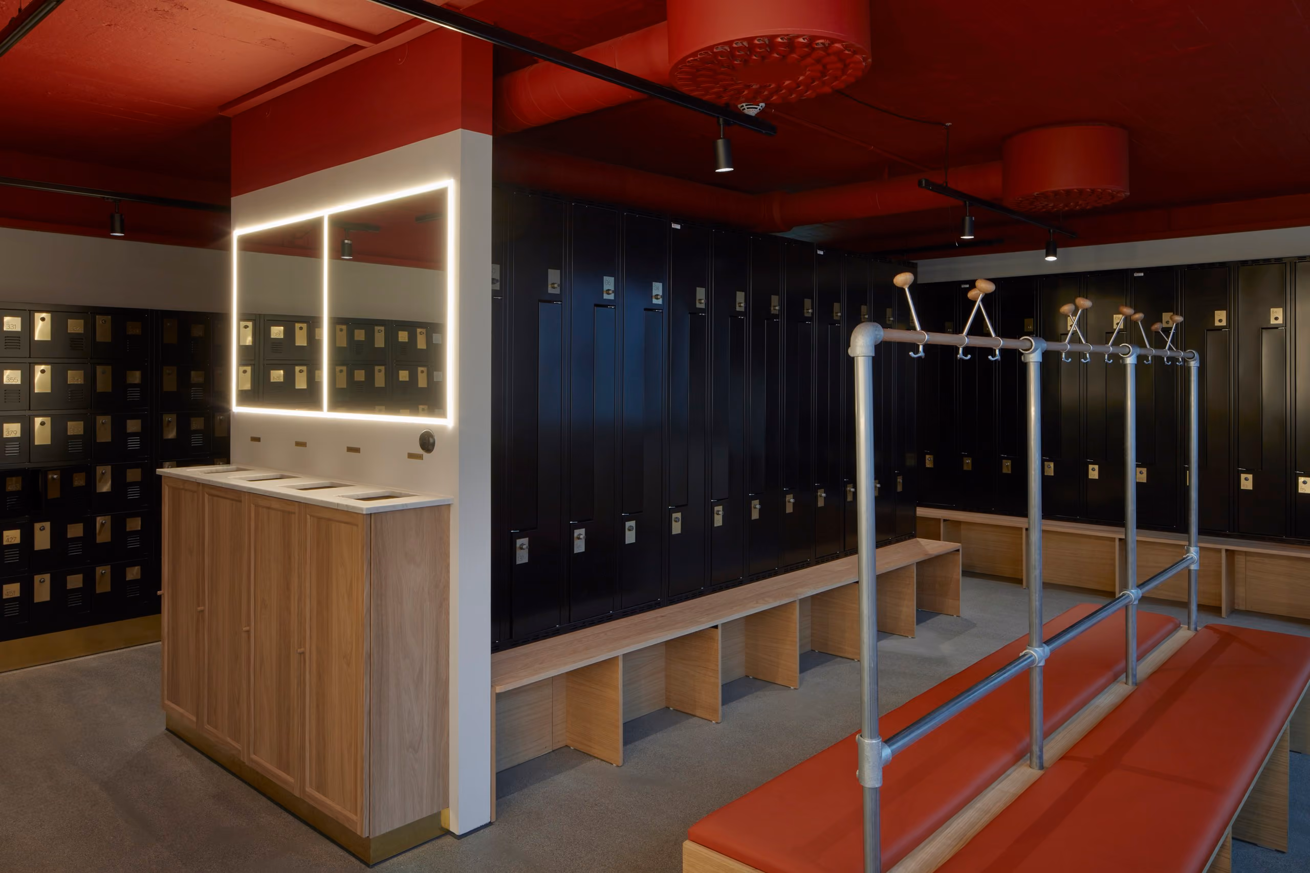 Changing room with dark lockers, a timber bench, a red ceiling and illuminated mirror.