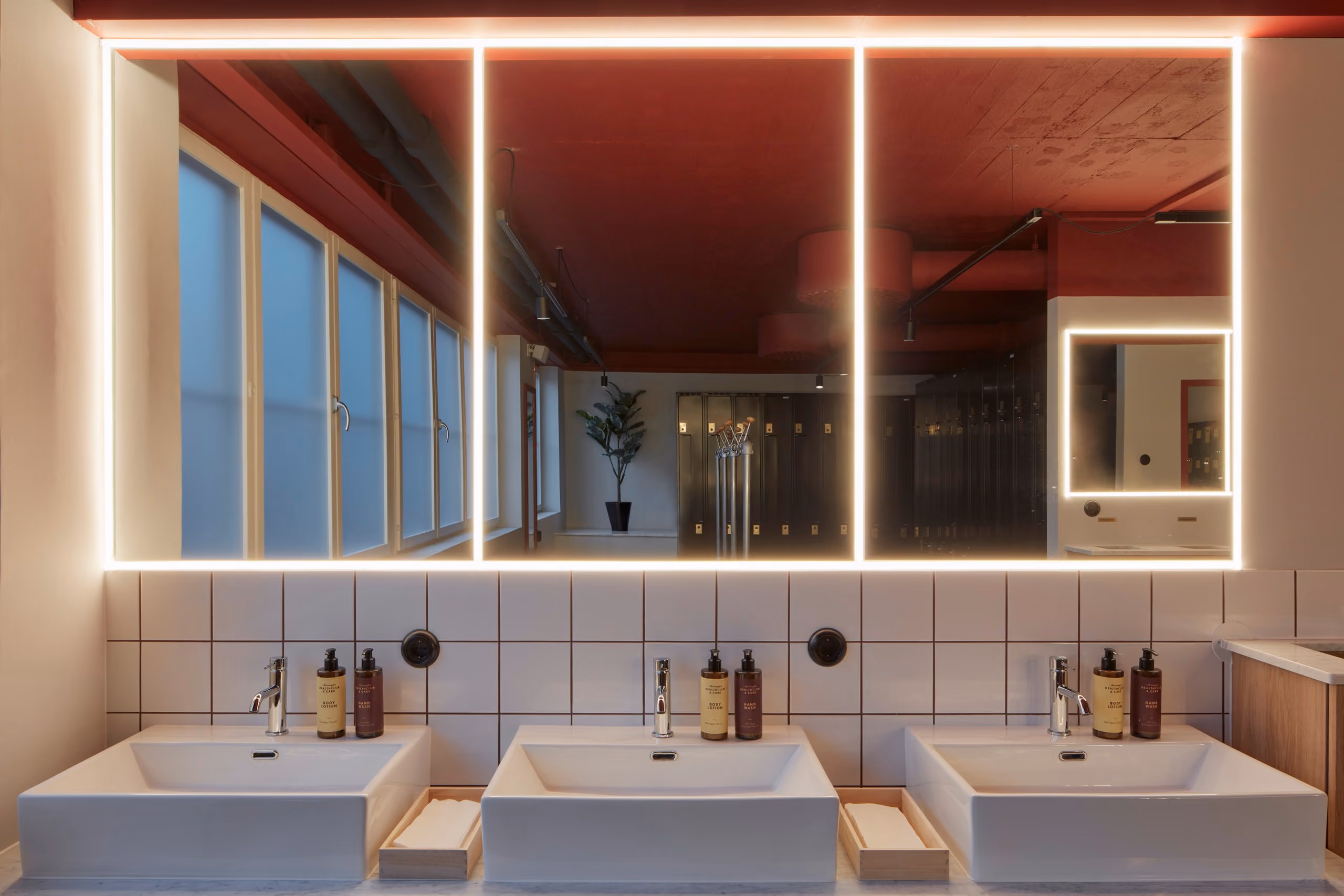 Three washbasins beneath large illuminated mirrors, with red ceilings reflected above white tiles.