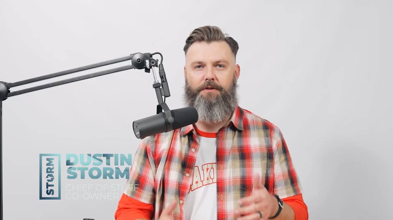 Bearded man with styled hair wearing a red and beige plaid shirt speaking into a studio microphone against a plain white background.