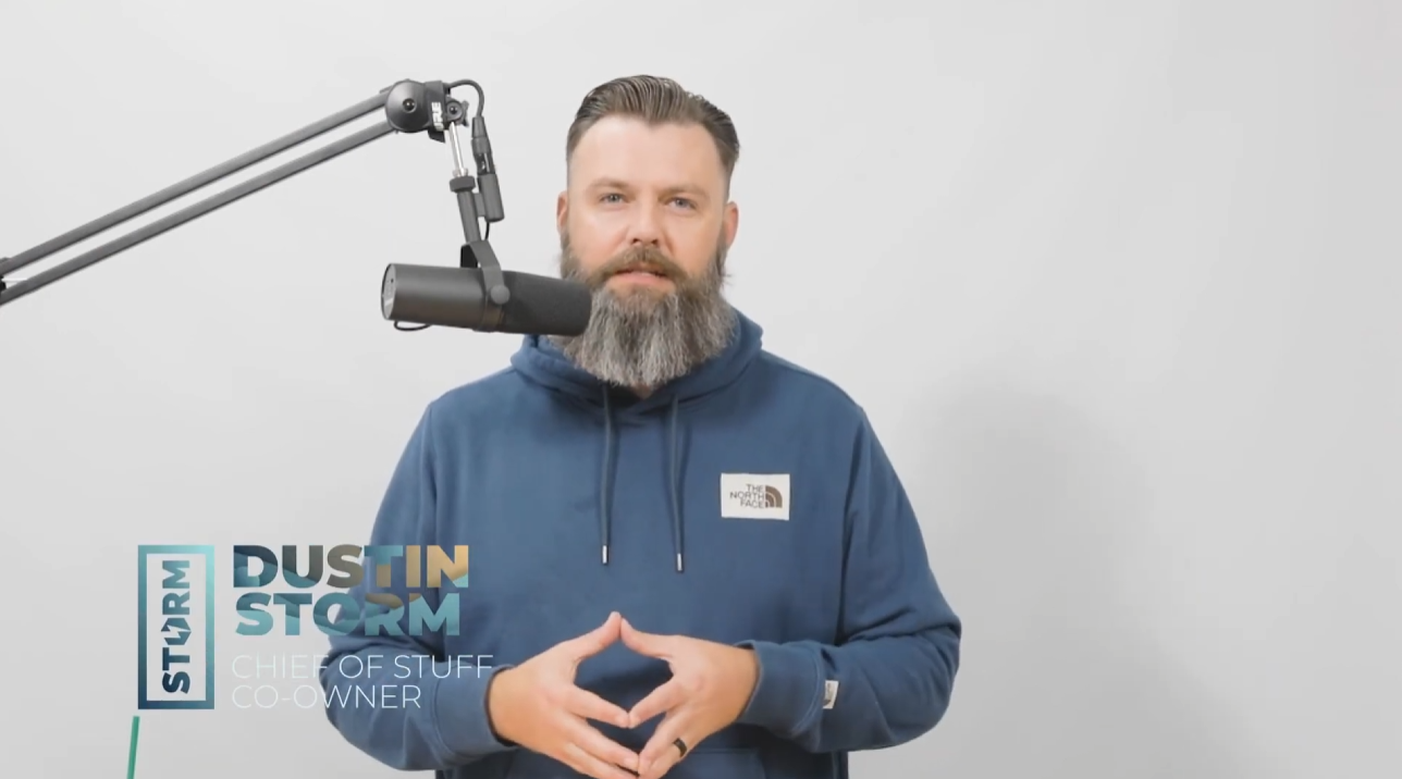 Man with a beard wearing a blue The North Face hoodie, speaking into a suspended microphone against a plain white background, with the text 'Dustin Storm Chief of Stuff Co-Owner' on the left.