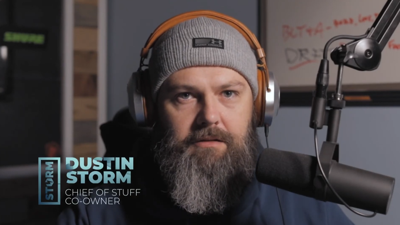 Man with a gray beard wearing headphones and a gray knit beanie speaking into a microphone.