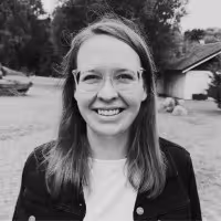 Smiling woman with glasses and long hair standing outdoors near a house.