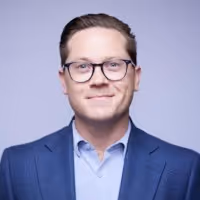 Man with glasses wearing a blue suit jacket and light blue shirt against a light purple background.