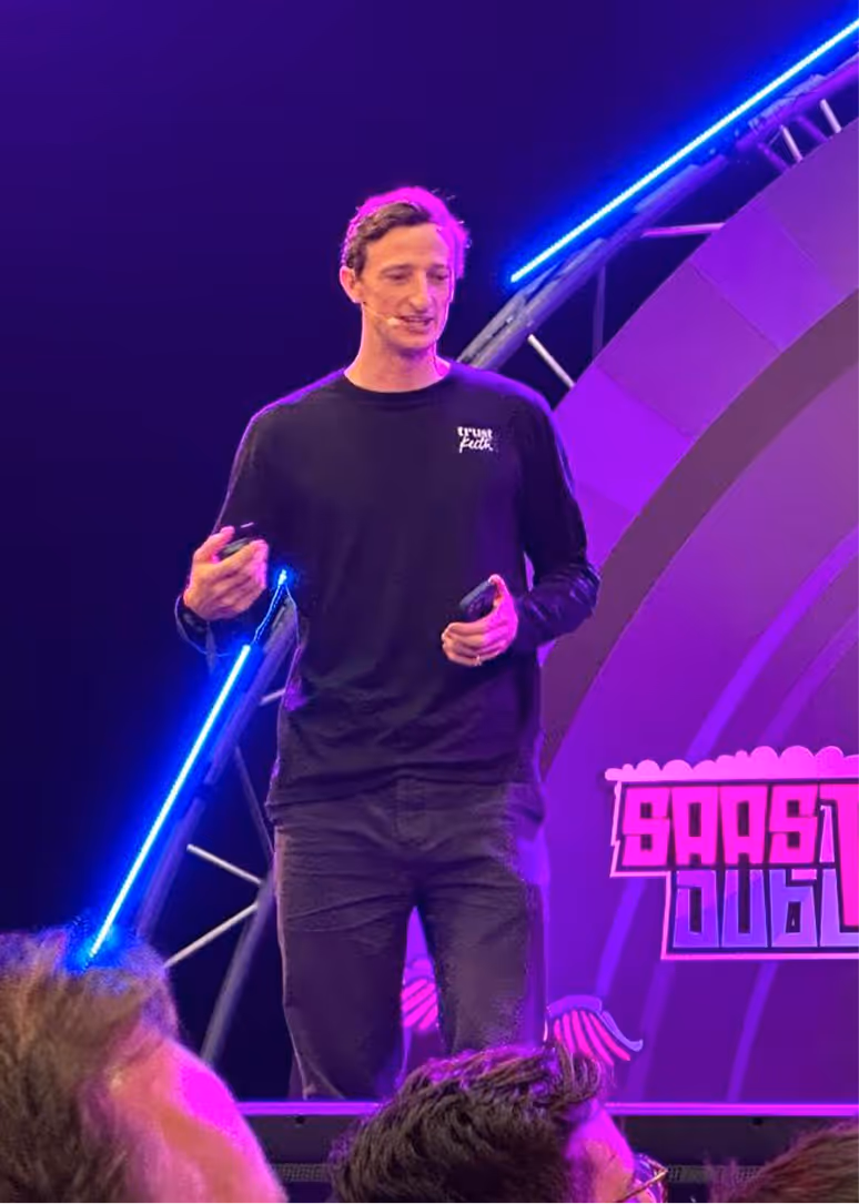 Man wearing a black long-sleeve shirt speaking on stage with blue and purple lighting, holding a phone.