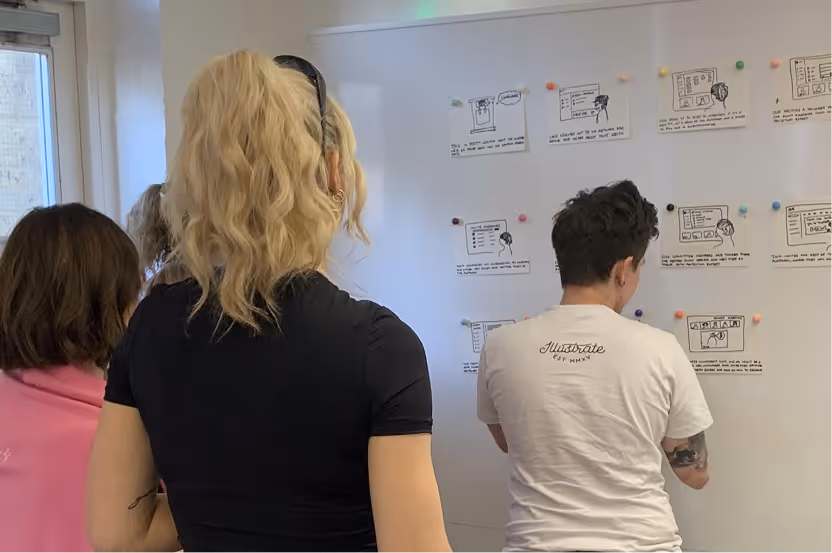 Three people stand facing a whiteboard wall with pinned sketches and notes in an office setting.