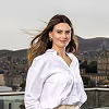 Smiling woman with long hair wearing a white blouse standing outdoors with a cityscape and hills in the background.