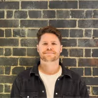 Smiling man with short hair and beard wearing a black jacket and white shirt standing in front of a dark brick wall.