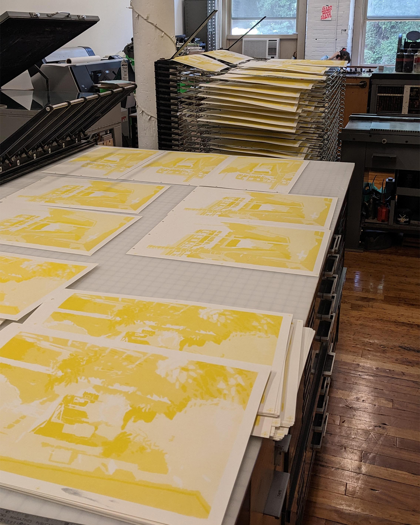 Stacks of yellow-tinted printed artworks laid out on a flat surface and drying racks in a printmaking studio.