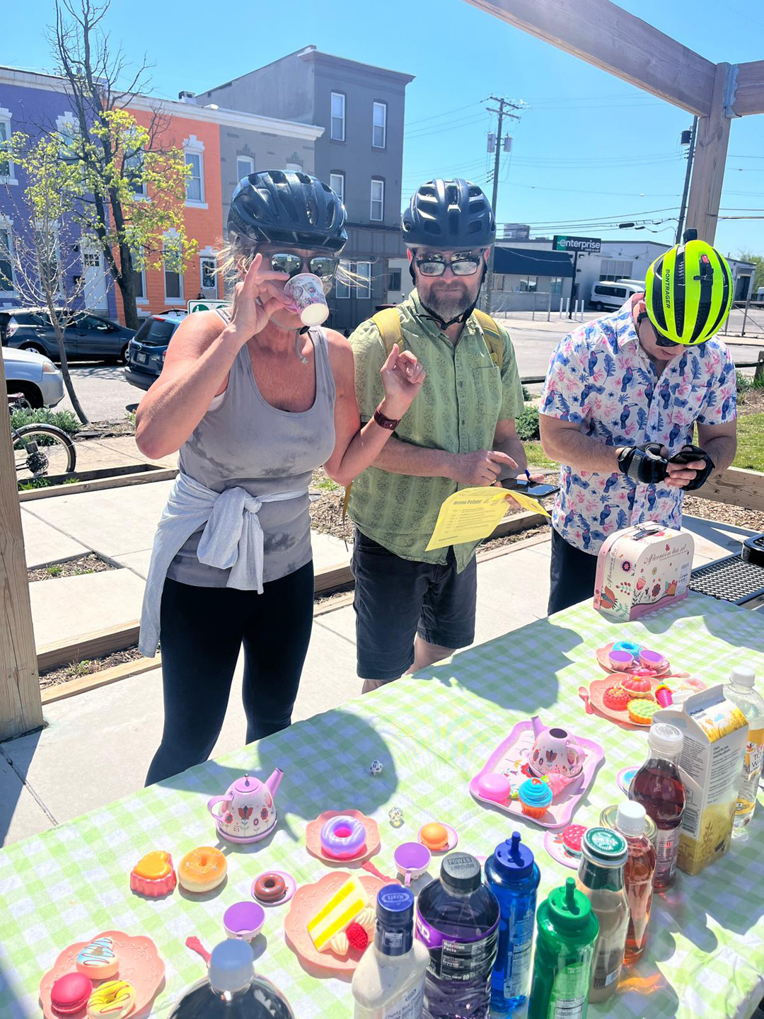 Three adults wearing bicycle helmets standing outdoors at a table with toy tea set and bottles on a sunny day.