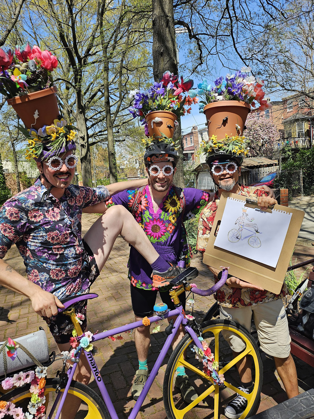 Three men wearing colorful floral shirts and glasses with flower frames, each with a flower pot hat holding artificial flowers, posing with a decorated purple bicycle, one holding a drawing of a person on a bike.