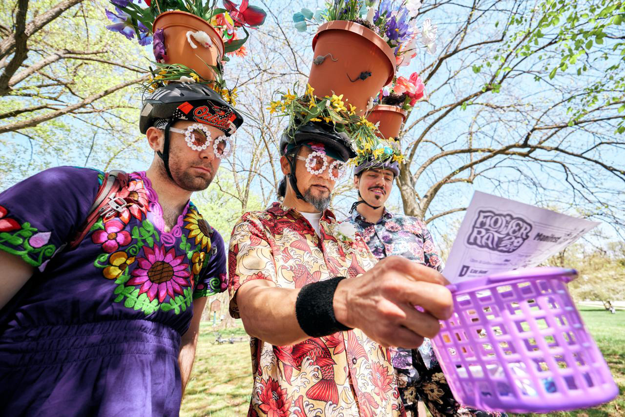 Three men wearing floral shirts, flower pot hats with fake mice, and flower-shaped glasses, holding a purple basket with a Flower Rave flyer in a sunny park.