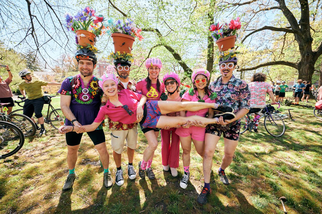 Group of cyclists in colorful outfits, two wearing flower pots on helmets, holding a woman in pink cycling clothes horizontally in a park on a sunny day.
