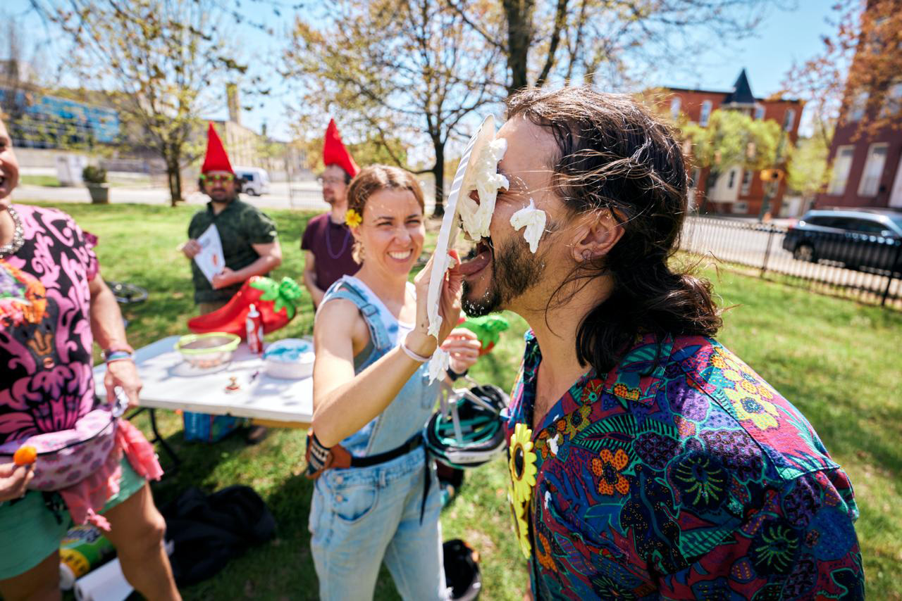 Woman in denim overalls playfully smearing whipped cream on a man's face outdoors during a sunny day, with people and a table in the background.