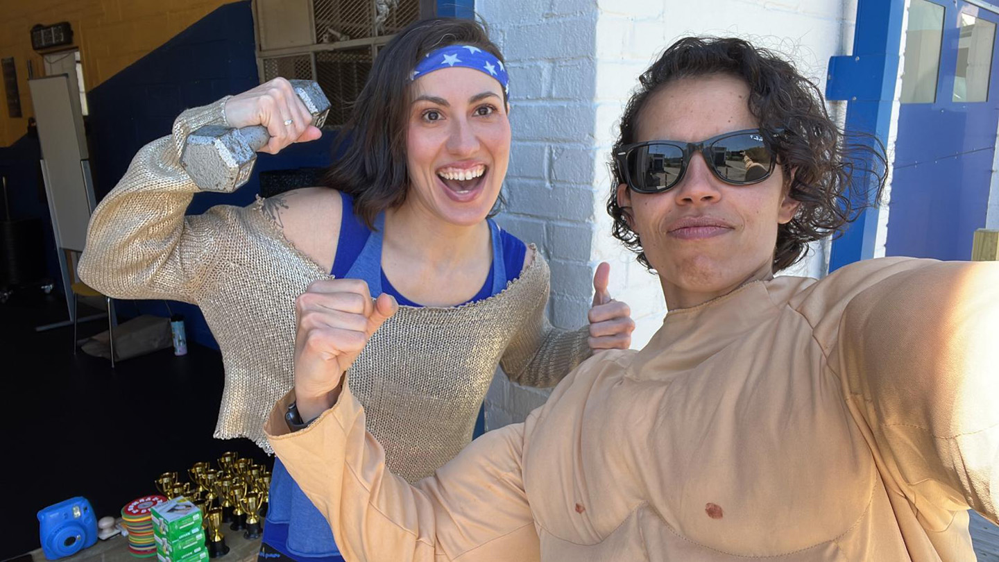 Two people posing for a selfie; one wearing a muscle costume and sunglasses making a thumbs-up, the other holding a dumbbell and smiling broadly with a star-patterned headband.