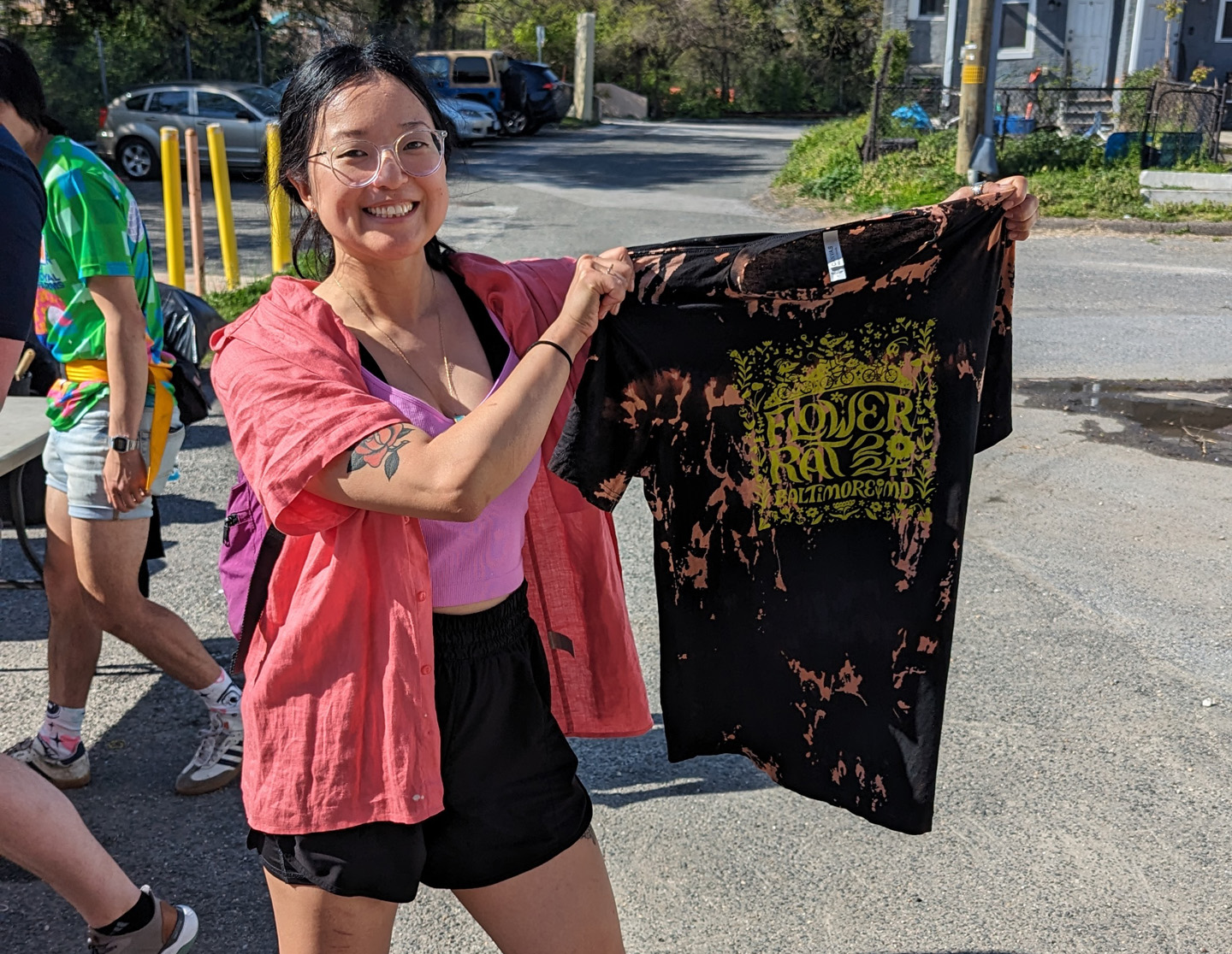 Smiling woman wearing glasses and a pink shirt holds up a black tie-dye t-shirt with yellow text and graphics reading 'Flower Rat Baltimore MD'.