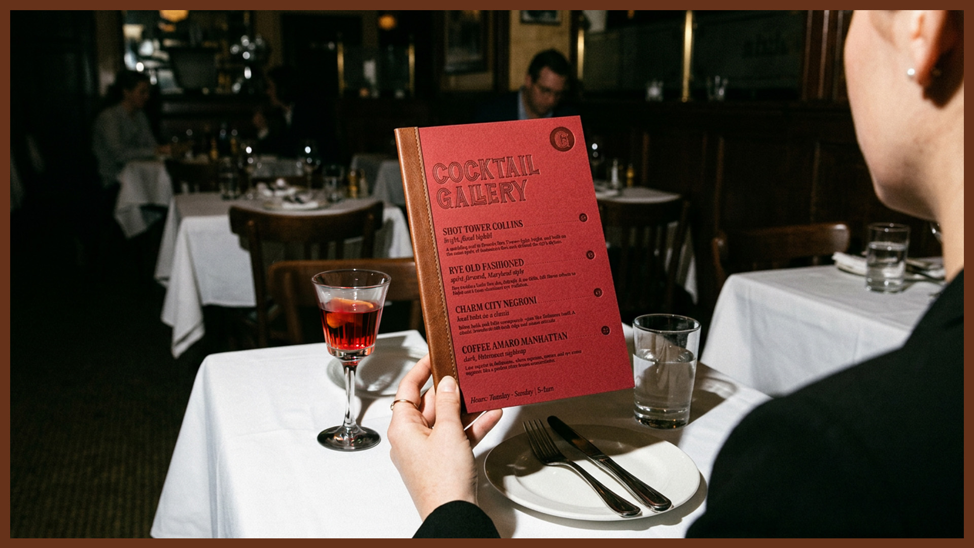 Person holding a red cocktail menu titled 'Cocktail Gallery' at a restaurant table with a glass of red cocktail, water glass, and white plate with cutlery.