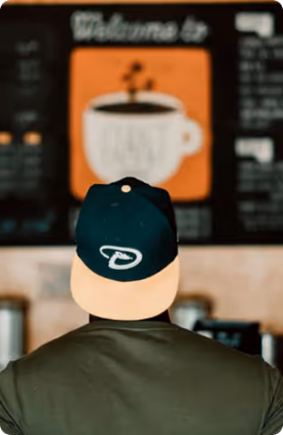 Person wearing a dark green and beige cap looking at a blurred café menu board with a coffee cup image.