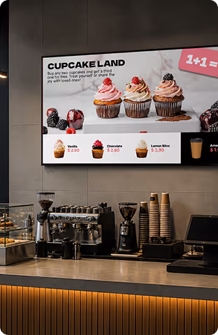 Bakery counter with coffee machines and a lit cupcake menu featuring four cupcake flavors and a 1+1 offer.