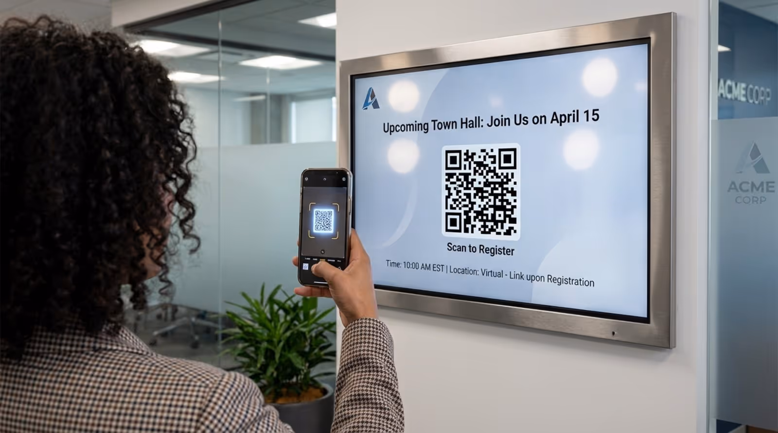 An employee scans a QR code on a digital bulletin board in a modern office hallway to register for a town hall.