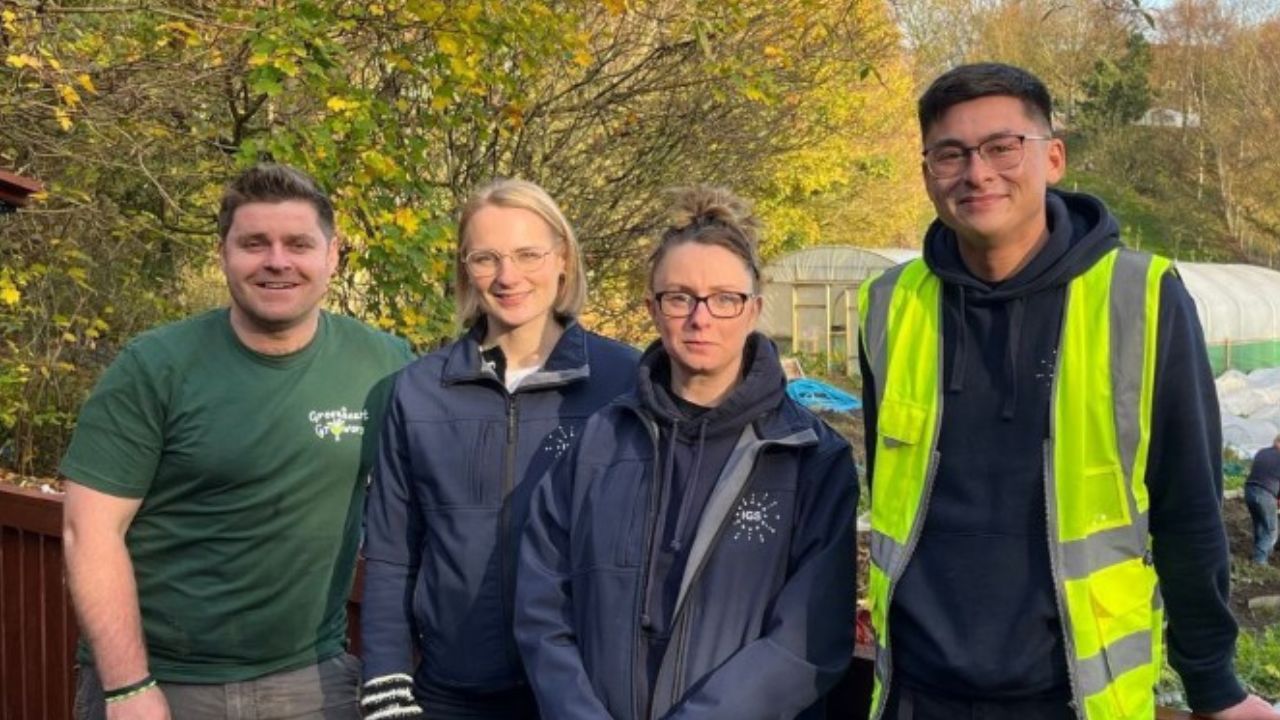 Group of four people standing outdoors in front of a lush, green garden with autumn foliage. One person is wearing a green t-shirt, two are in navy jackets, and one is in a high-visibility vest. The background features trees and a greenhouse.