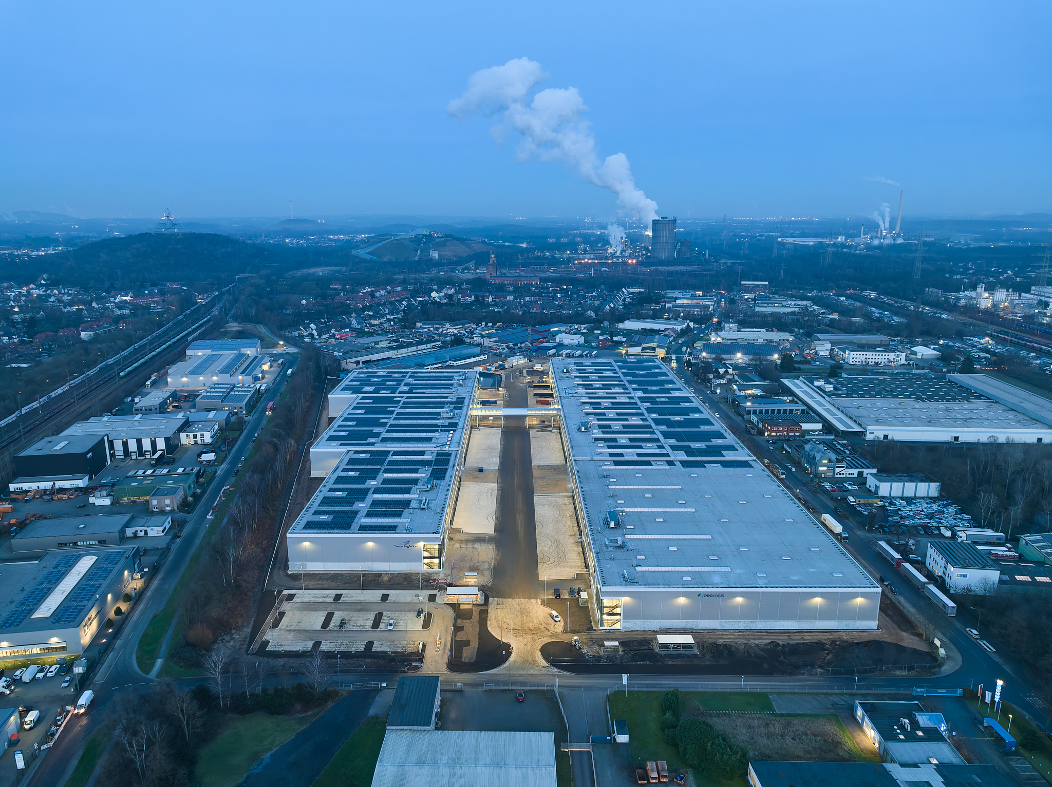 Drohnenaufnahme in der blauen Stunde, leuchtende Hallen im Abendlicht.