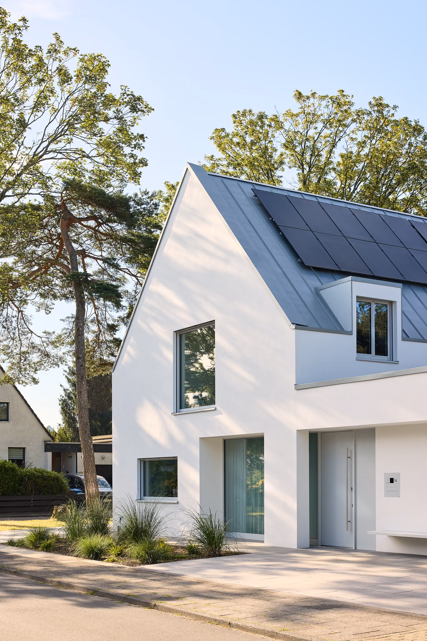 Modern weißes Haus mit Solarpanelen auf dem Dach und gepflegtem Vorgarten an sonnigem Tag.