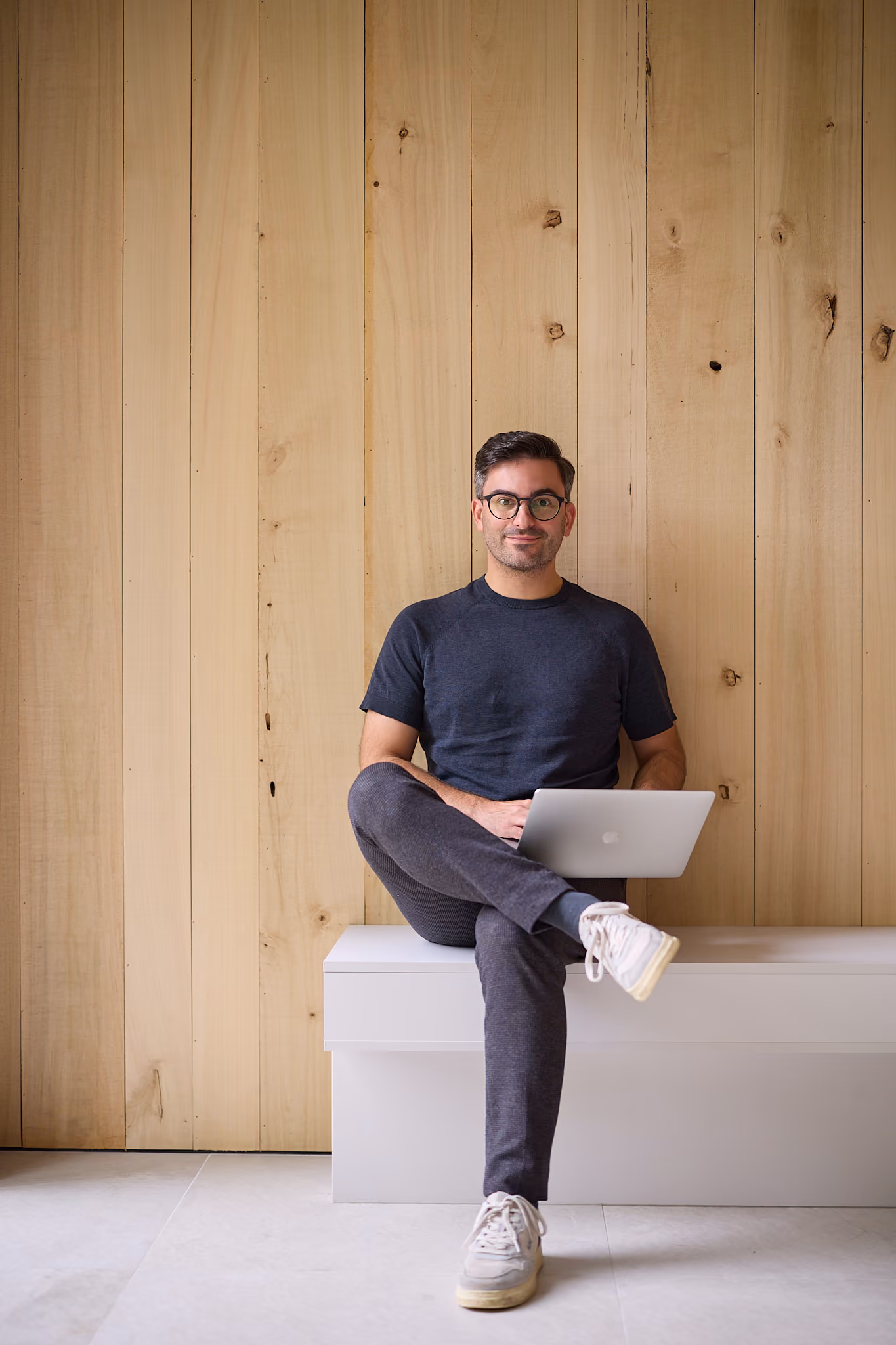 Mann mit Brille sitzt auf Bank vor Holzwand und arbeitet mit Laptop auf dem Schoß.
