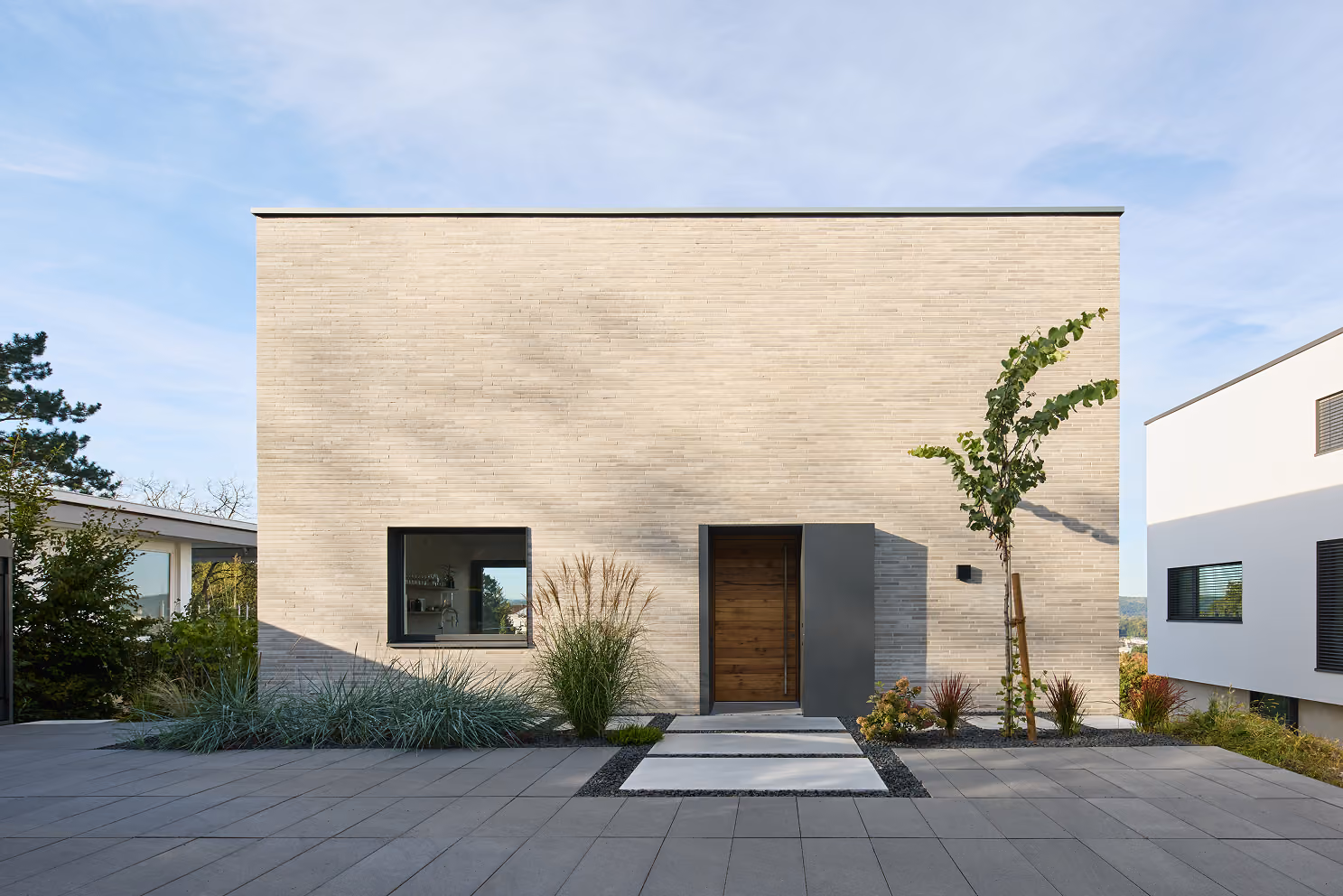 Modern minimalist house front with beige brick, wooden door, square window, and landscaped yard.