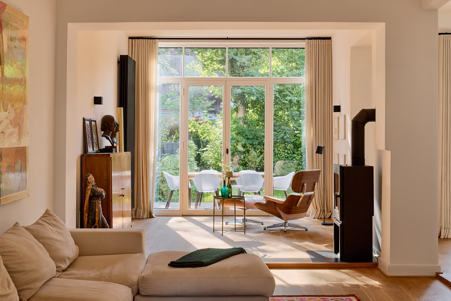 Wohnzimmer mit beige Sofa, Ledersessel, Holzkamin und großem Fenster mit Blick auf Garten.