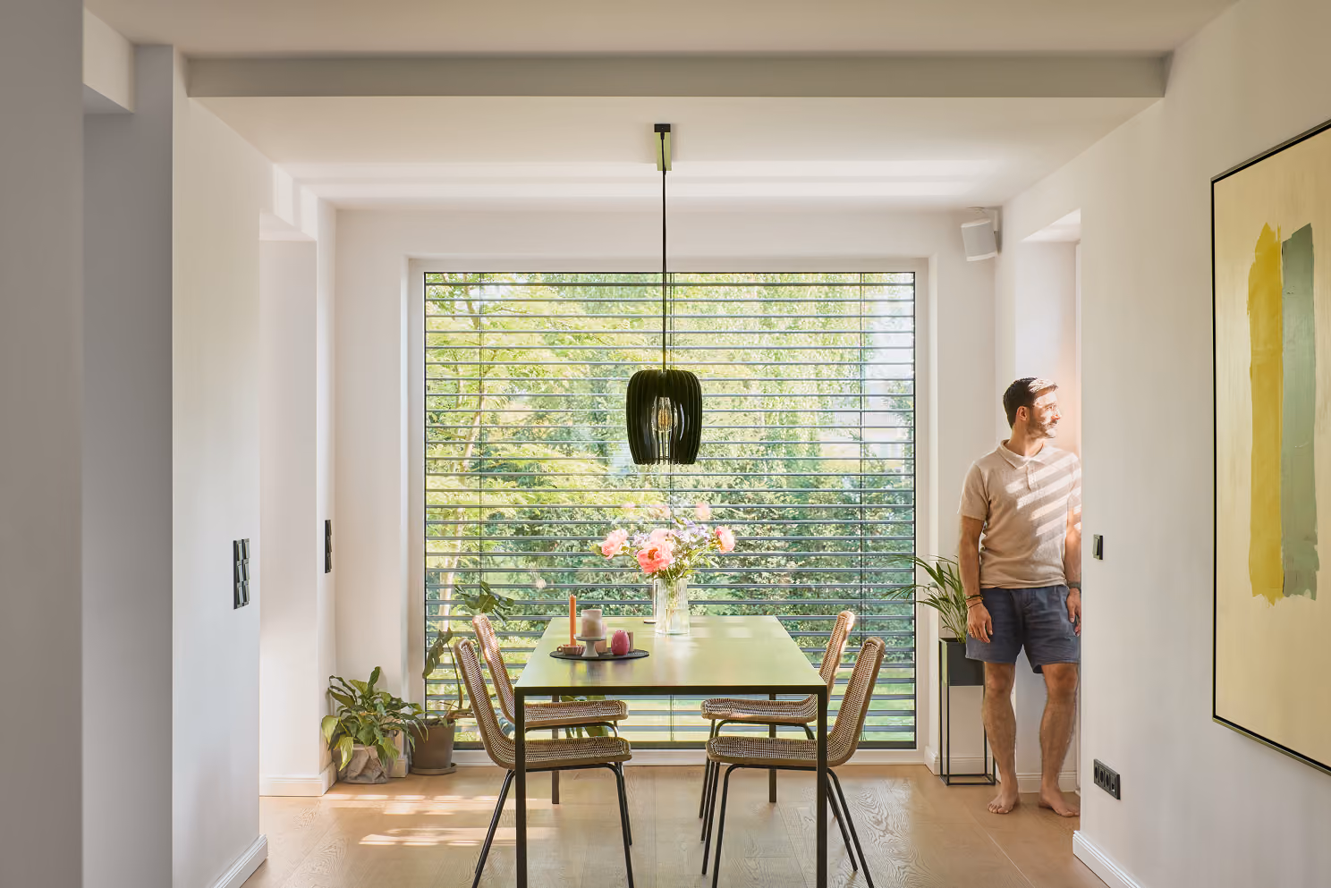 Esszimmer mit großem Fenster, Tisch mit Blumen, Stühlen und einem Mann, der nach draußen schaut.