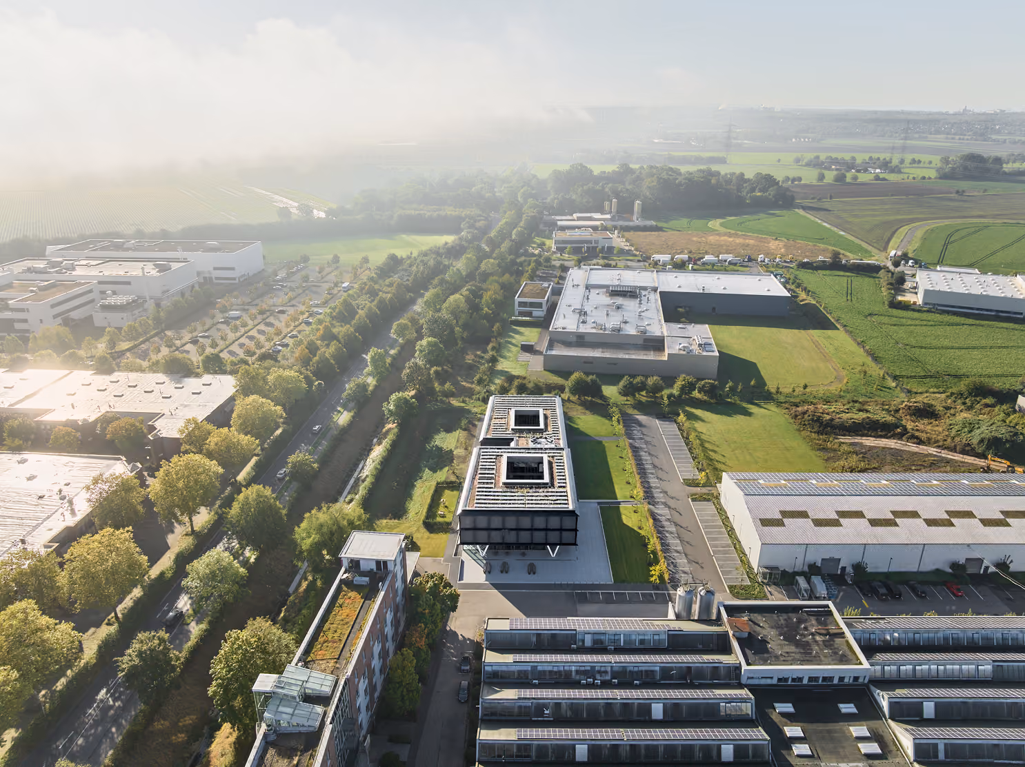 Luftaufnahme eines Büro- und Industriegebiets mit grünen Feldern und bewaldeten Straßen.