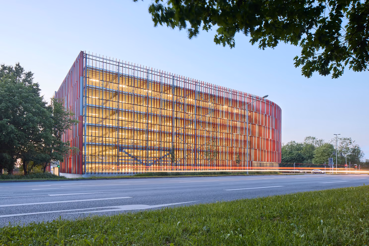 Parkhaus mit roter Fassadenverkleidung und beleuchtetem Innenraum an einer Straße bei Dämmerung.