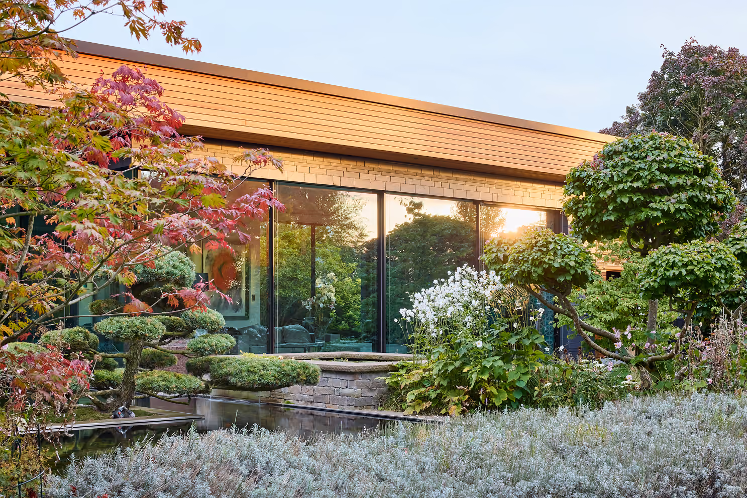 Moderne Glasfassade eines Hauses mit gepflegtem Garten und verzweigten Zierbäumen bei Sonnenuntergang.