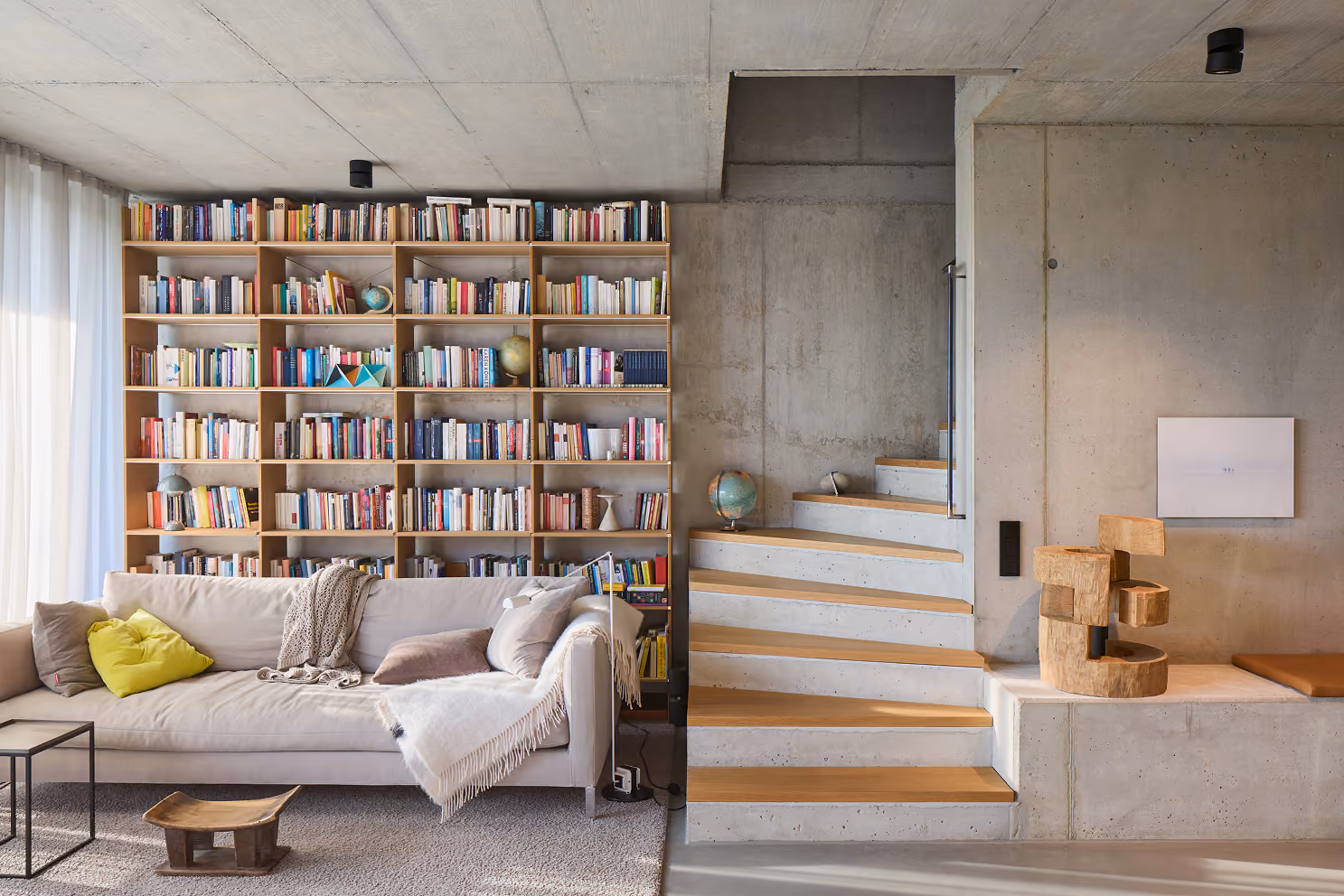 Wohnzimmer mit großem Bücherregal, Sofa mit Kissen, Treppe aus Holz und Beton, und moderner Skulptur.