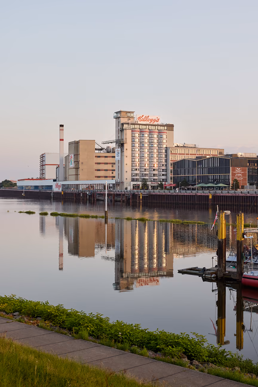 Kellogg's Fabrik am ruhigen Fluss mit Spiegelungen, Gehweg und Bootssteg im Vordergrund.