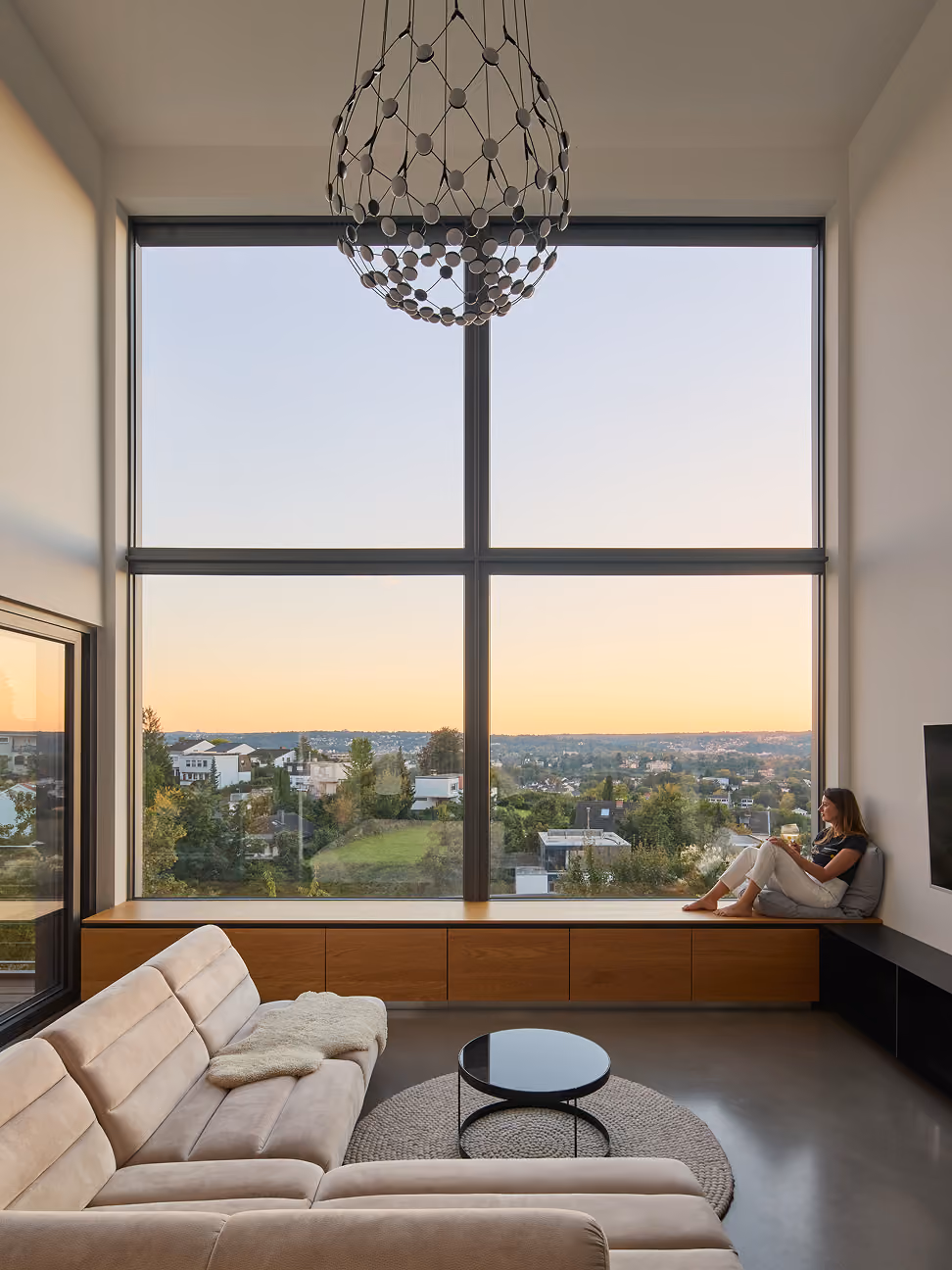 Frau sitzt auf Fensterbank mit Aussicht auf Häuser und Landschaft bei Sonnenuntergang.