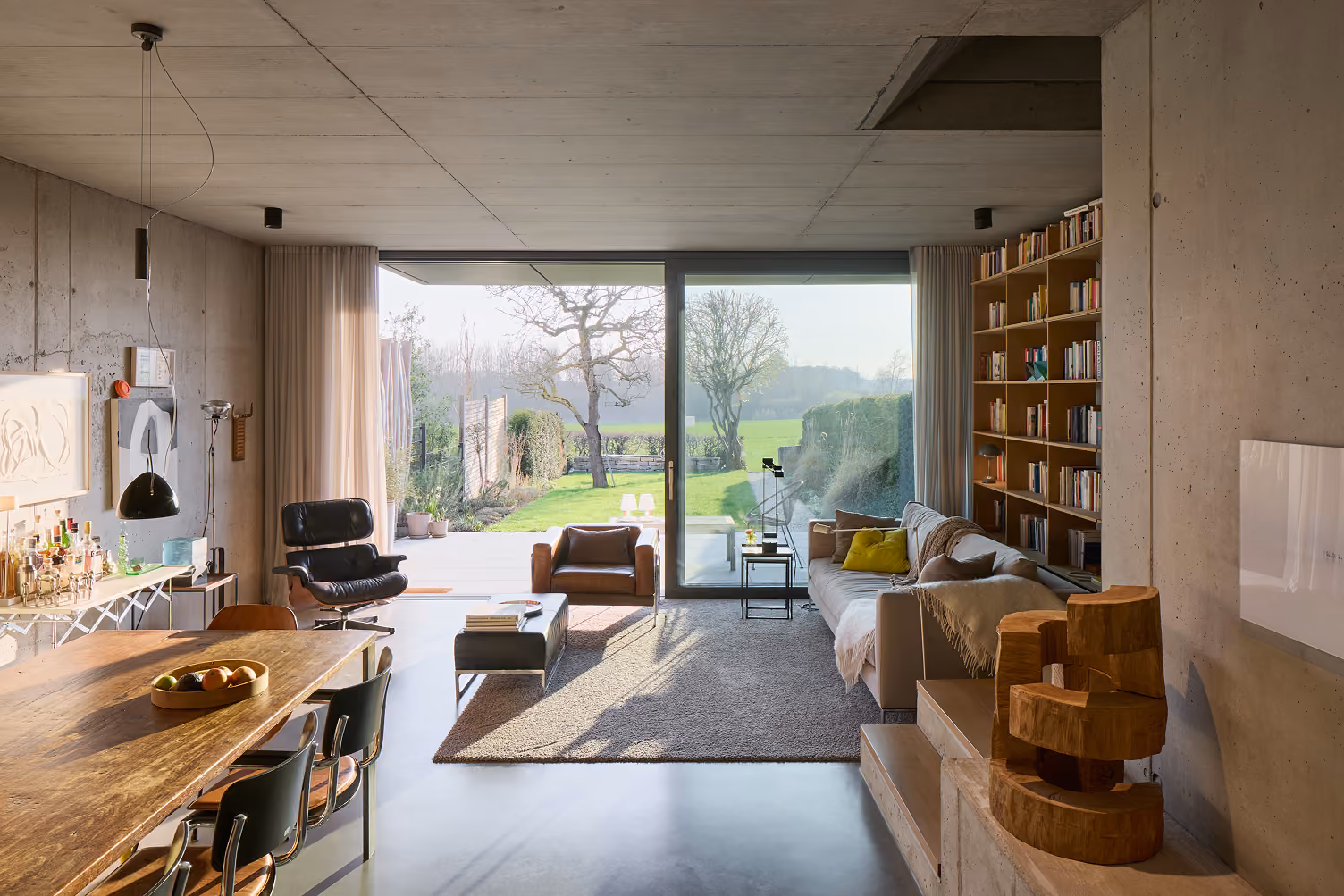 Modernes Wohnzimmer mit großem Fenster, Sofa, Ledersessel, Bücherregal und Blick auf Garten.