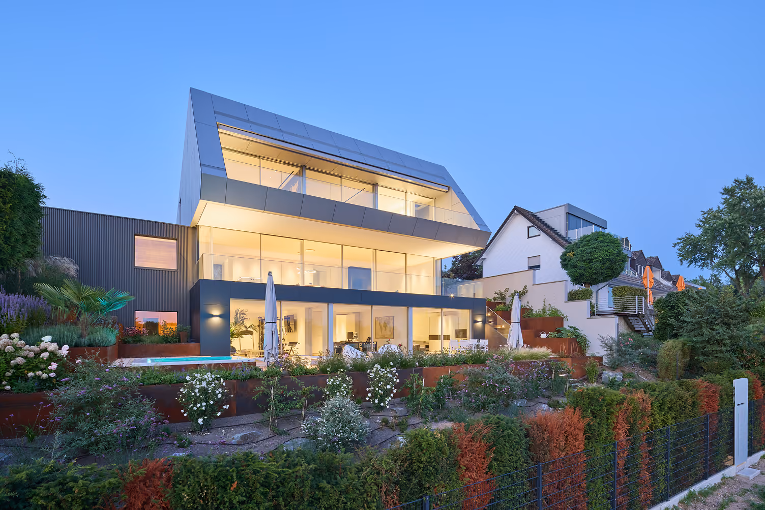 Modern hell beleuchtetes Haus mit großen Fenstern, Garten und Nachbarhäusern bei Dämmerung.