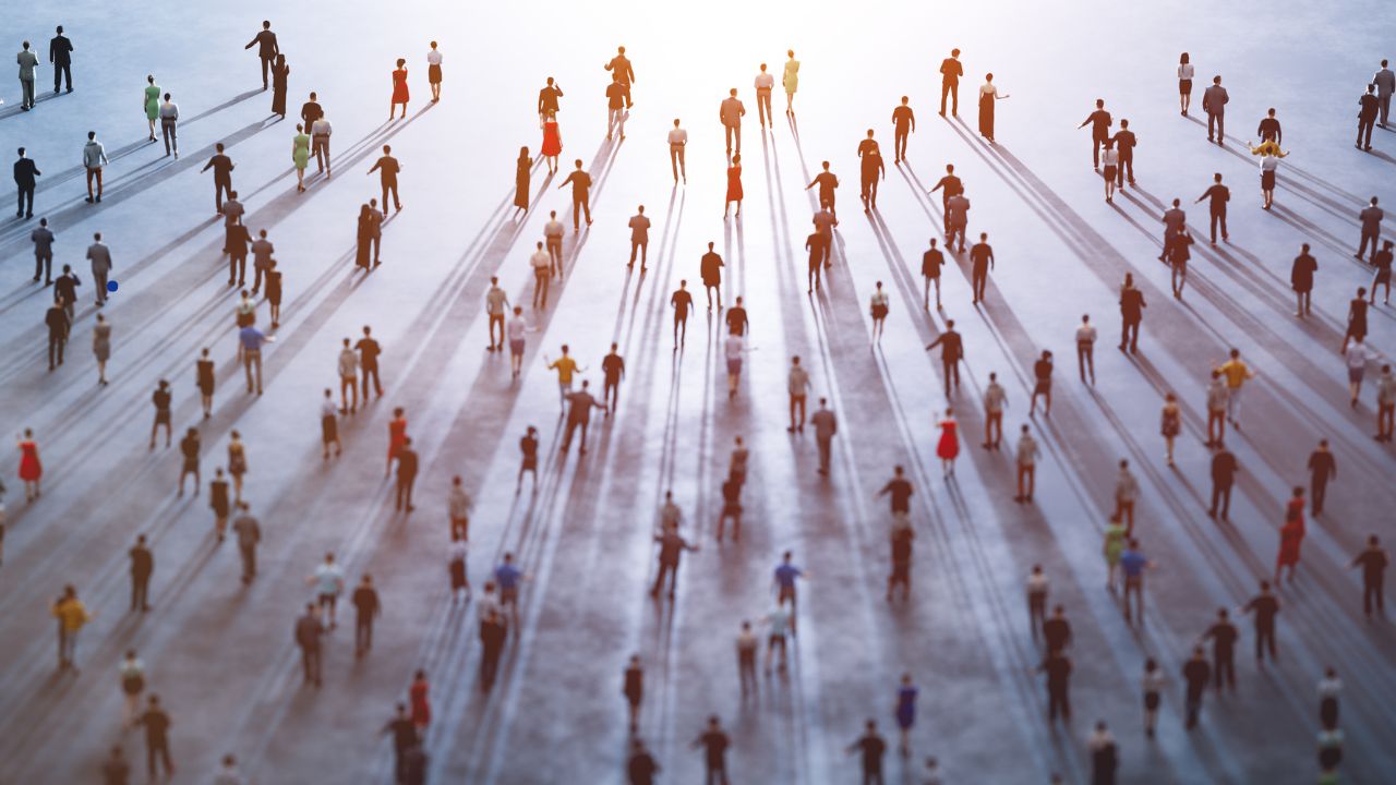 Una pequeña bandera de España sobre una base dorada situada en el extremo izquierdo de un escritorio de madera clara. Al fondo, se observa un portalápices con bolígrafos, un cuaderno abierto y una ventana iluminada que da al exterior.