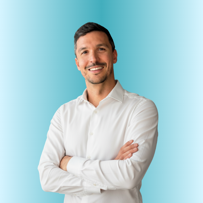 Retrato de medio cuerpo de un hombre con cabello oscuro corto y barba ligera, vestido con una camisa blanca formal. Se muestra con los brazos cruzados y una sonrisa amplia y cercana frente a un fondo gris oscuro degradado.