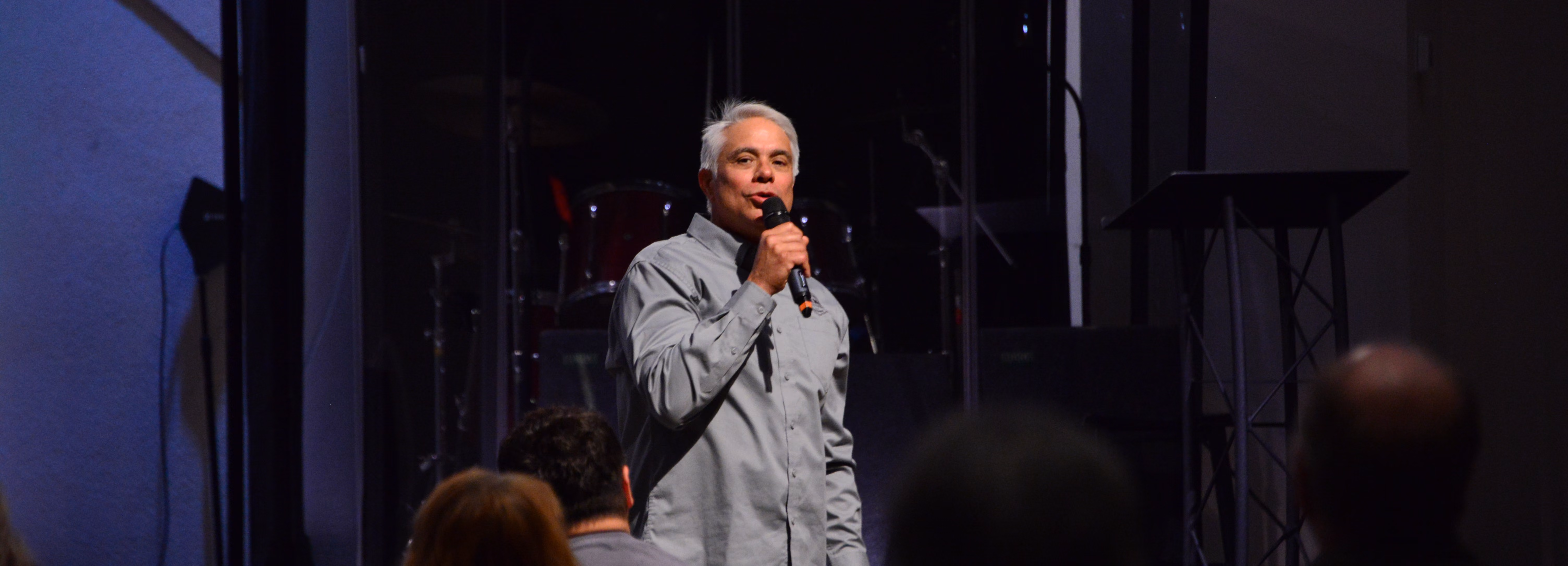 Man with gray hair and a gray button-up shirt speaking into a microphone on a dimly lit stage with silhouettes of audience members in the foreground.