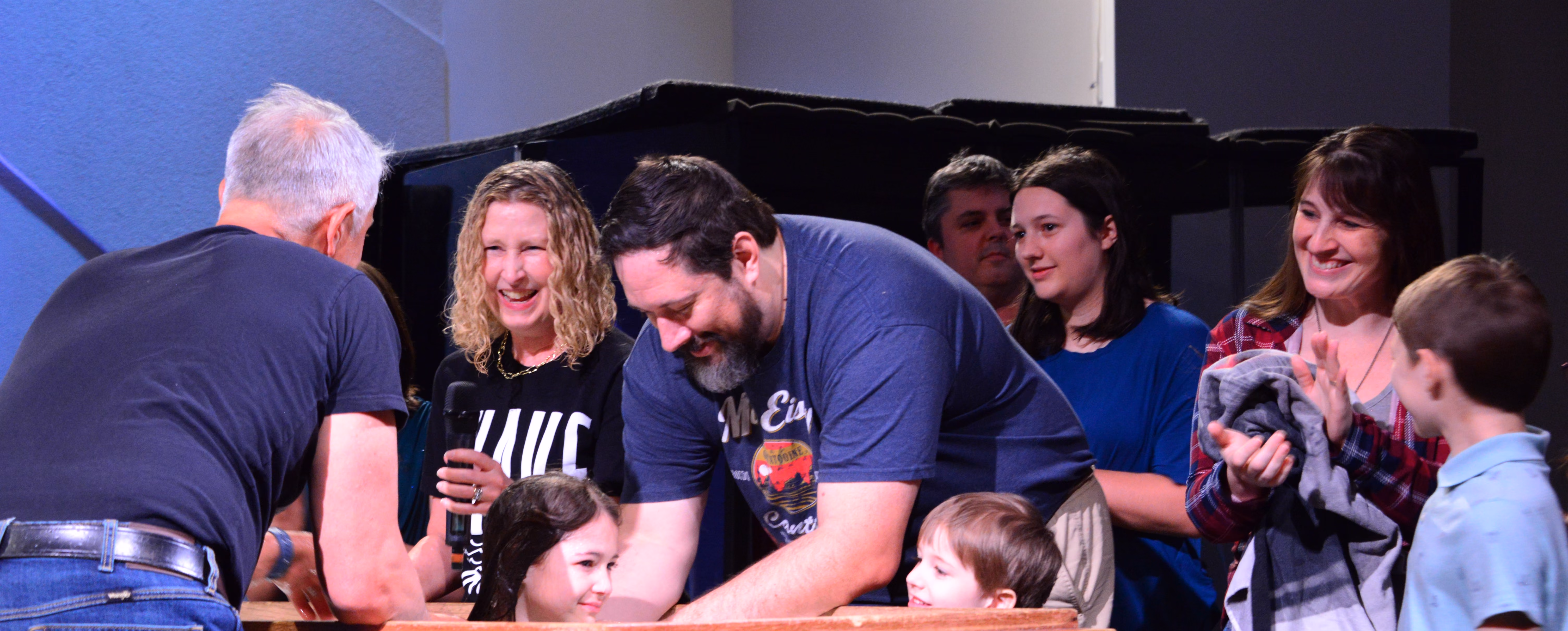 A group of adults and children smiling and interacting closely, with some people holding blankets, in an indoor setting with a black background.