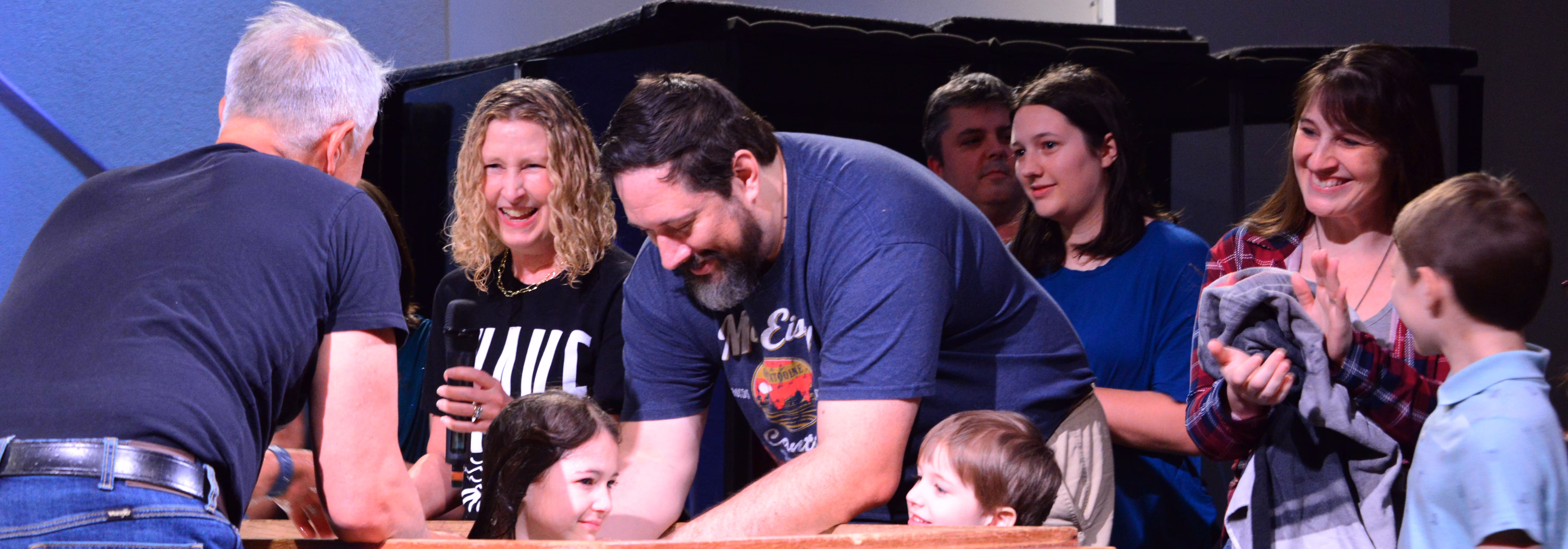 Group of adults and children gathered around a baptismal pool during a baptism ceremony indoors, some smiling and holding towels.