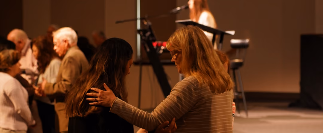 Two women in a church setting, one with her hand on the other woman's shoulder in a gesture of comfort or prayer, with other people and a person speaking at a microphone in the background.