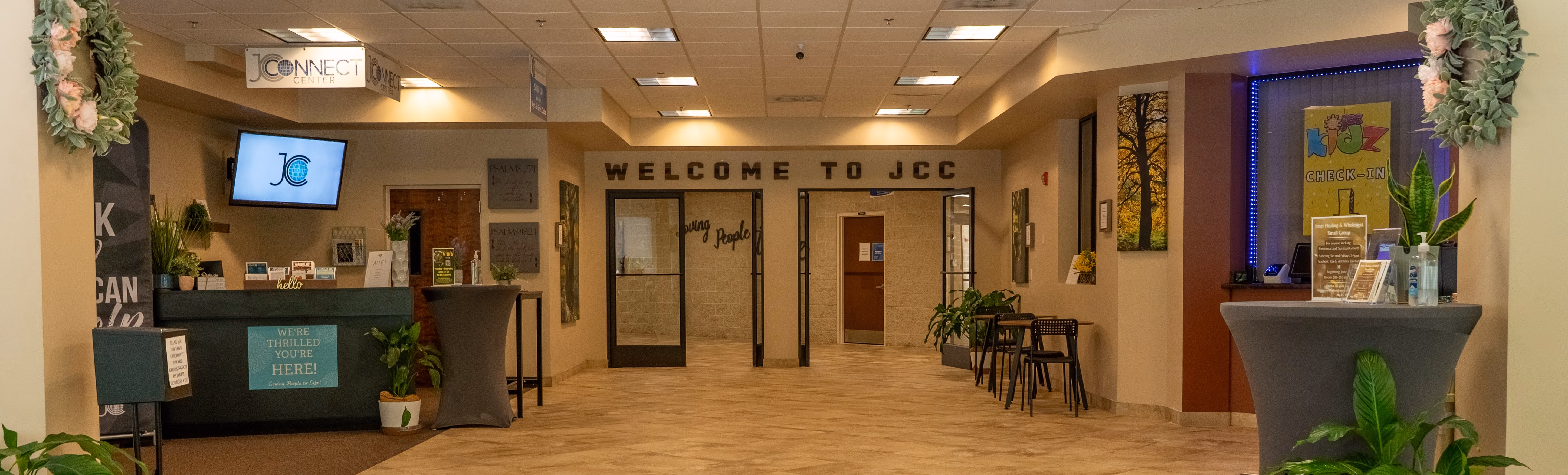 Interior entrance area with 'WELCOME TO JCC' sign above glass doors, reception desk on the left, and tables with plants on both sides in a well-lit hallway.