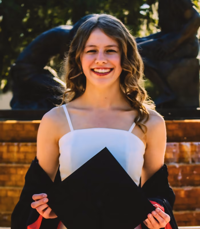 Young woman with wavy hair in a white sleeveless top smiling and holding a black graduation cap in front of her against a background of trees and a brick wall.