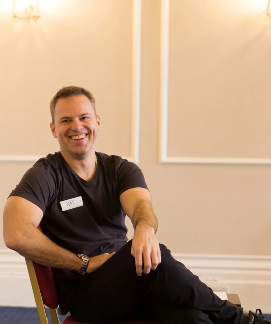 Nat Sharratt wearing a black t-shirt and sitting relaxed on a red chair in a beige room.