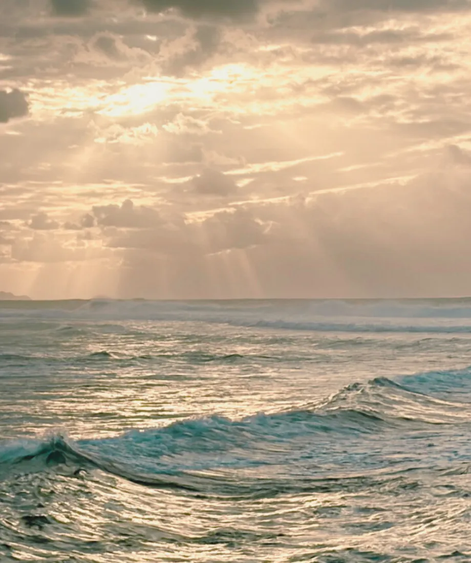 Sun rays shining through clouds over a calm ocean with gentle waves.